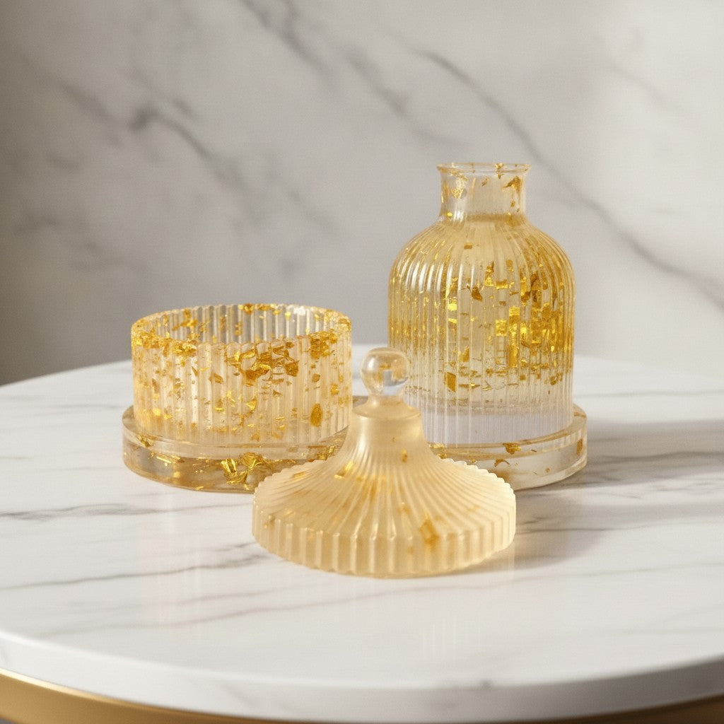 Gifts Under 20 GBP - A decorative ribbed resin storage set with a small bottle, a round container, and a matching lid, all in a translucent pale gold tone with embedded metallic gold flakes, displayed on a white marble surface against a marble wall.