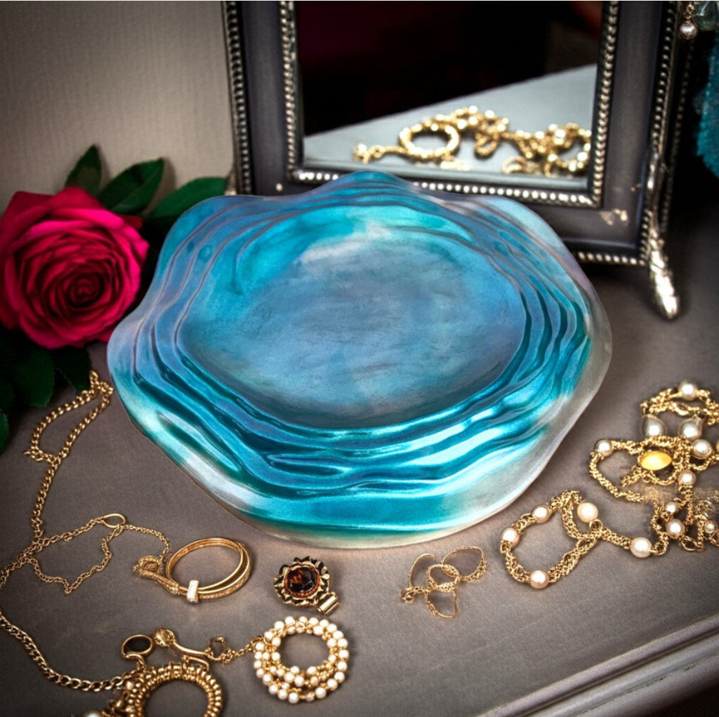 Decorative Trays - A round, organic-shaped resin decorative tray with a wavy, layered pattern in shades of blue and teal, styled on a grey surface surrounded by gold jewellery, a red rose, and a mirror.