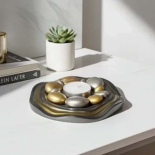 A decorative Candle Holder Plate holding a white tea light candle, surrounded by smooth gold and silver stones, displayed on a white tabletop with a small potted succulent and books in the background.