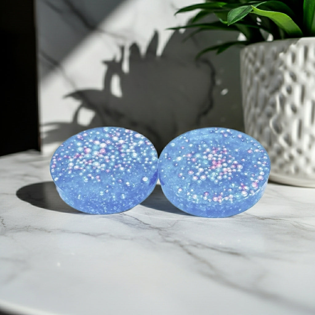Two thick round blue glittery coasters with iridescent beads embedded, resting on a white marble surface next to a potted plant.