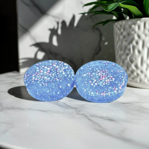 Two thick round blue glittery coasters with iridescent beads embedded, resting on a white marble surface next to a potted plant.