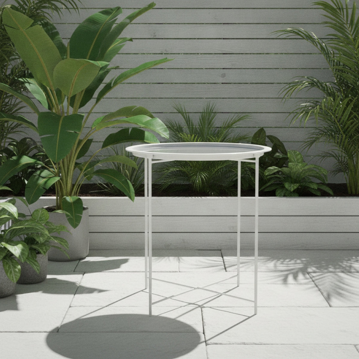 A white metal round side table with a thin frame and a clear glass top, set on a light tiled patio surrounded by potted green plants against a gray wooden fence.