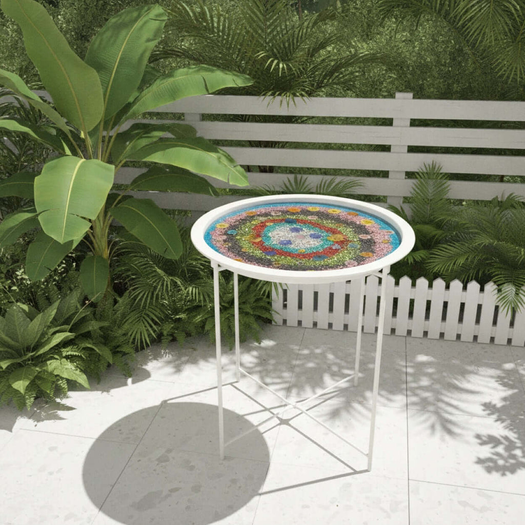 A round outdoor side table with a colorful mandala-style mosaic top and a white metal frame, set on a patio in front of a white fence and green plants.