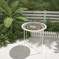 A round outdoor side table with a colorful mandala-style mosaic top and a white metal frame, set on a patio in front of a white fence and green plants.