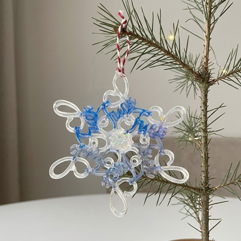 Novelty Christmas Tree Decorations - A clear and blue snowflake-shaped Christmas ornament with a red-and-white striped hanging loop, shown hanging from a pine tree branch indoors.