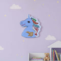 A light blue unicorn head wall hanging with glittery multicolor mane and tail details, mounted on a lavender wall with cloud and star decals above a small shelf with books and a teddy bear.