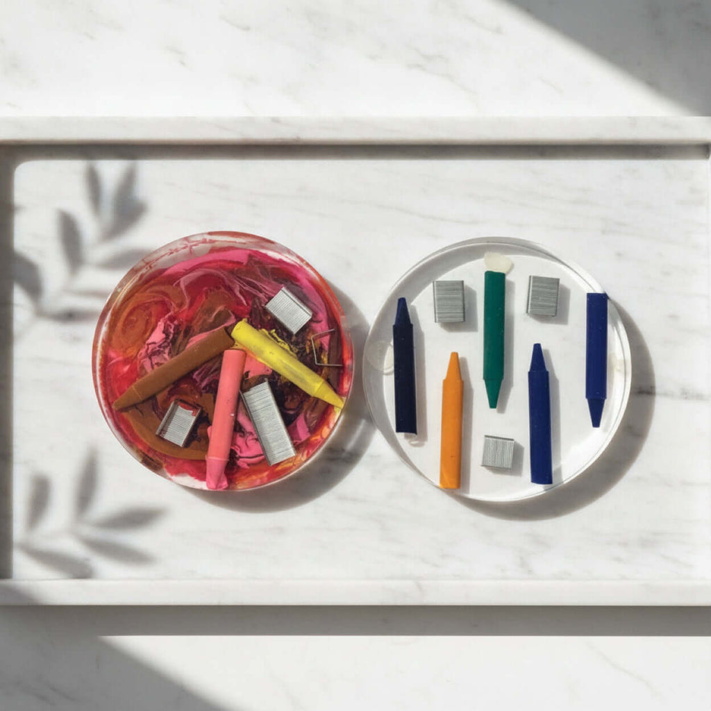 A styled flat-lay of two round coasters with crayons and staples embedded in resin. The left coaster has a pink swirl background with several crayons embedded and the right coaster has several loose crayons in blue, green, orange, and grey with staples embedded.