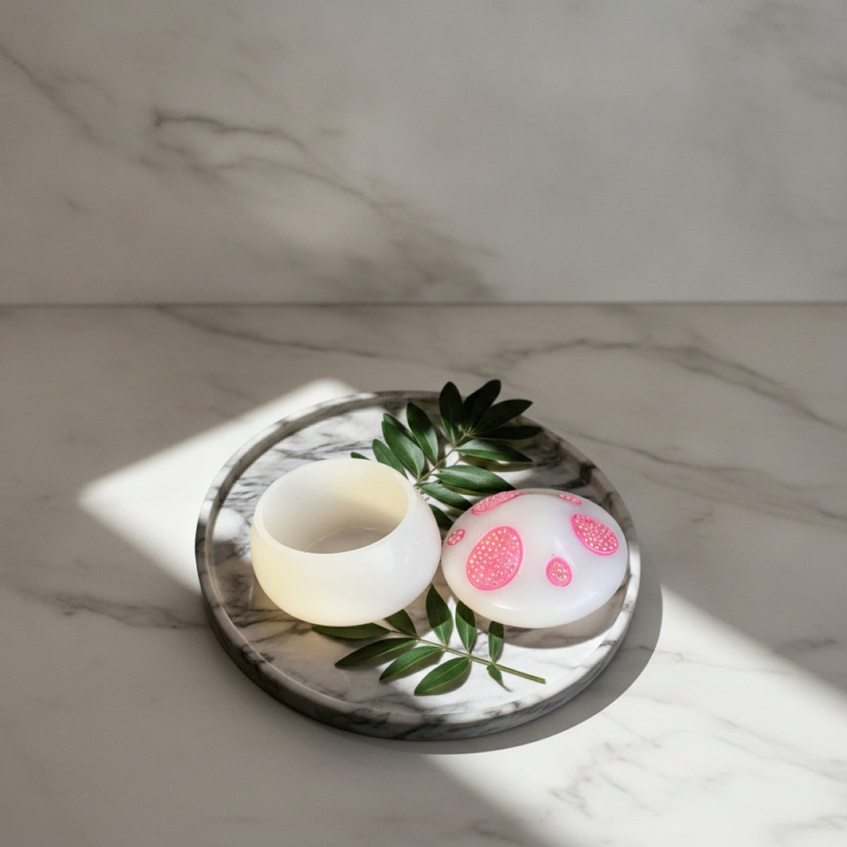 A small white resin container and matching round lid with pink polka dots sit on a round marble tray with green leaves, photographed on a marble surface with natural sunlight and shadows.