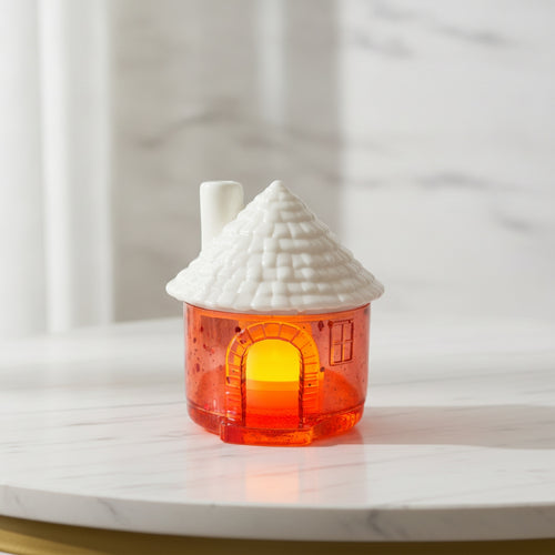 Candle Holder Houses - A house-shaped candle holder with a textured white ceramic roof and a translucent orange glass body glowing with warm light, set on a white marble surface.