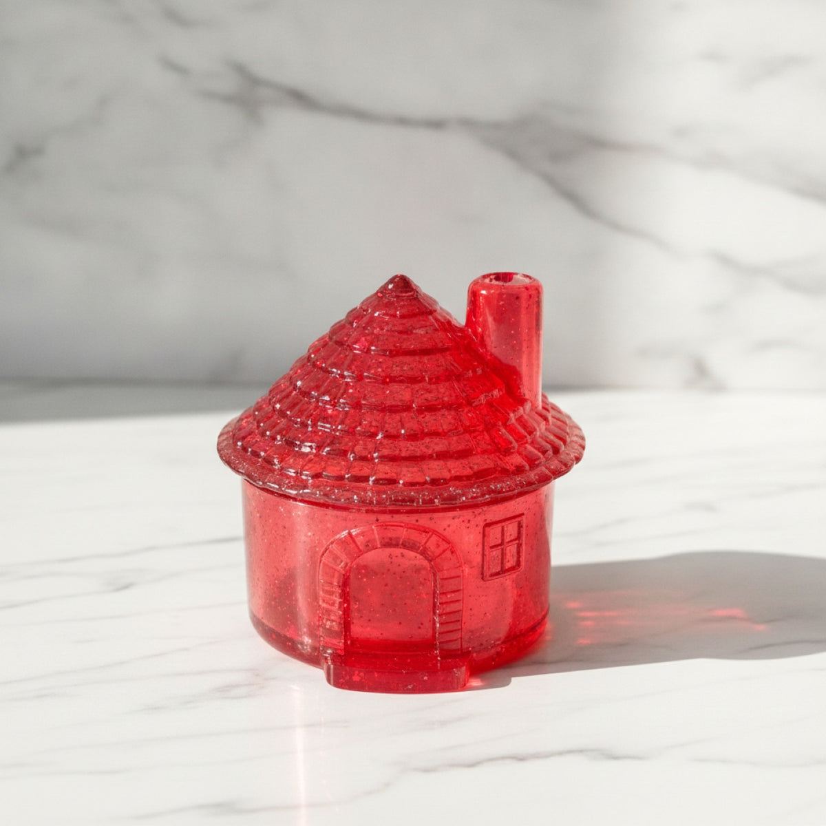 A small translucent red house-shaped object with a textured conical roof and chimney, photographed on a white marble surface with a matching marble background.