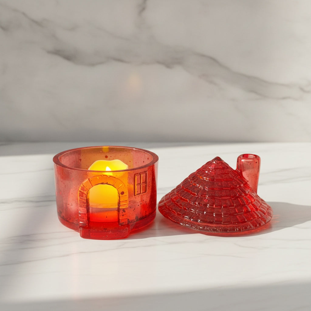 A red translucent house-shaped candle holder with a small lit yellow candle inside, shown next to its matching red textured lid on a white marble surface and background.