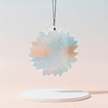 A sunflower suncatcher with a soft pastel gradient of light blue, pink, and mint green, suspended from a silver chain above a white round pedestal against a soft pink gradient background.