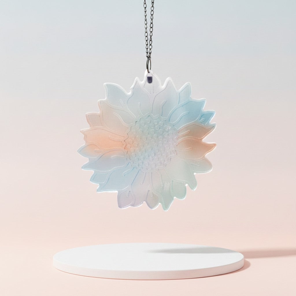 A flower-shaped sun catcher with a soft pastel gradient of light blue, pink, and mint green, suspended from a silver chain above a white round pedestal against a soft pink gradient background.