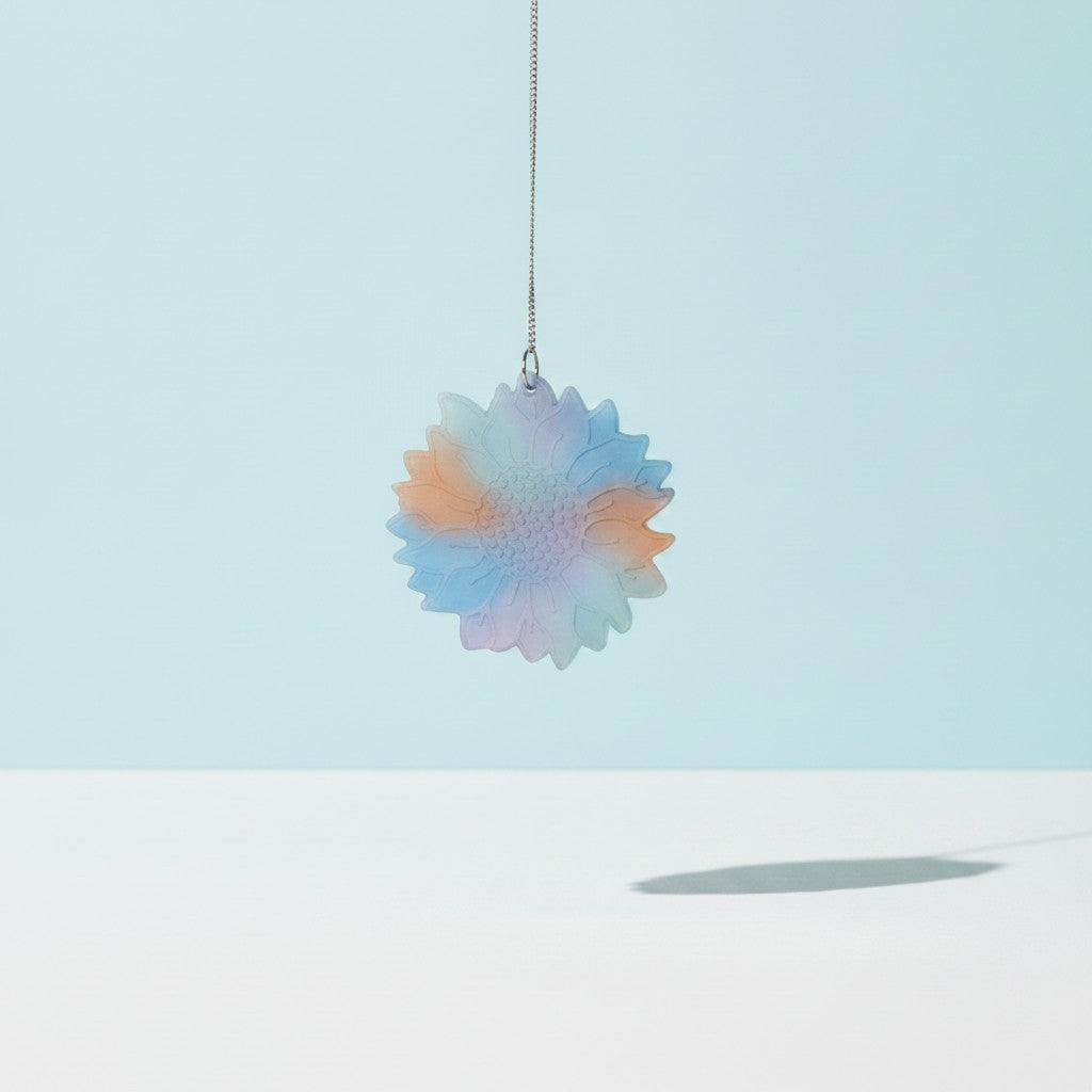 A flower-shaped sun catcher made of translucent resin with a pastel gradient of blue, pink, and green, hanging from a silver chain against a soft blue background