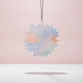 A flower-shaped sun catcher made of translucent resin with a pastel gradient of blue, pink, and green, hanging from a silver chain against a soft pink background.