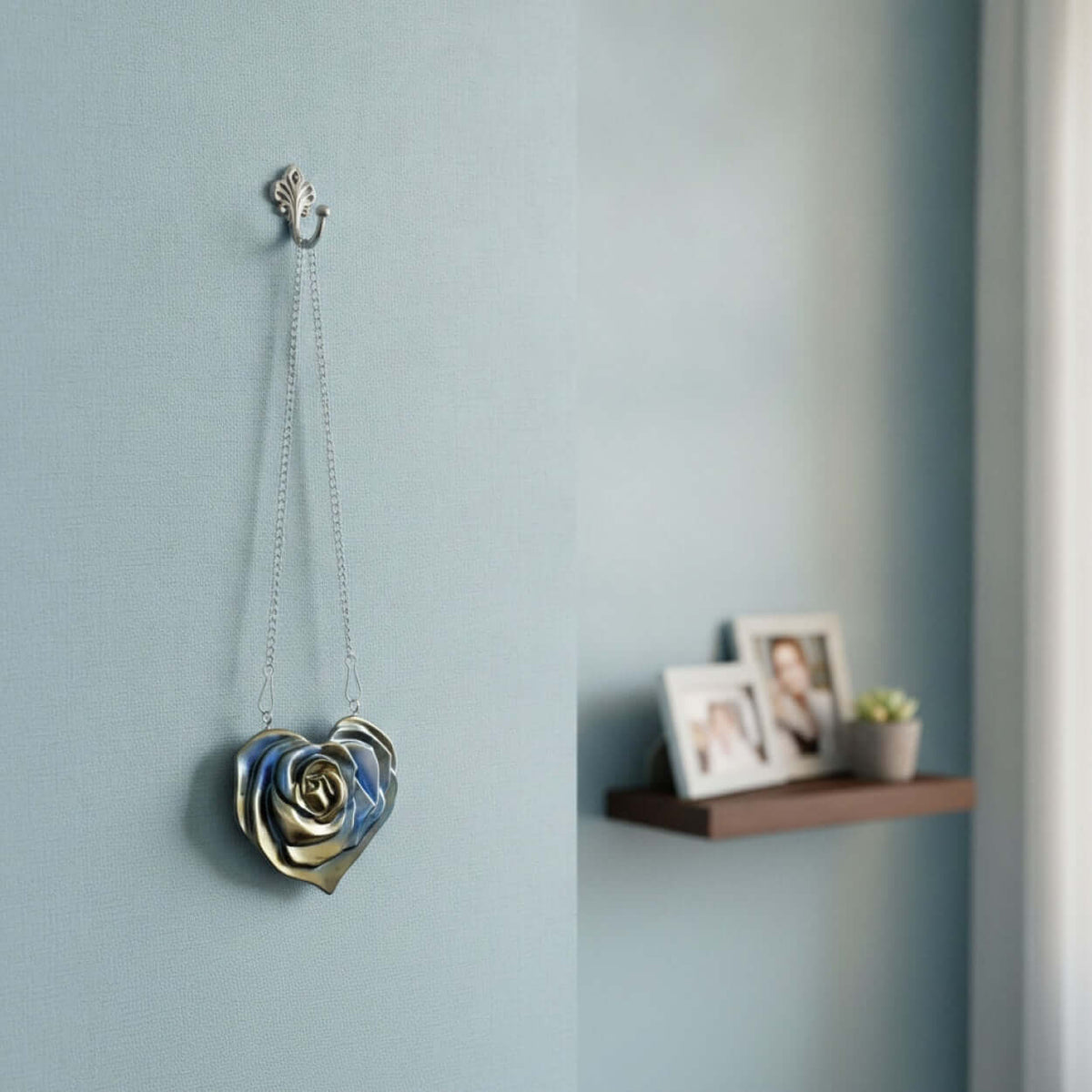 A metallic silver hook hanging on a pale blue wall, with a Heart Wall Decal rose-shaped ornament suspended from it by two chains. framed pictures and a small potted succulent are visible on a shelf to the right.