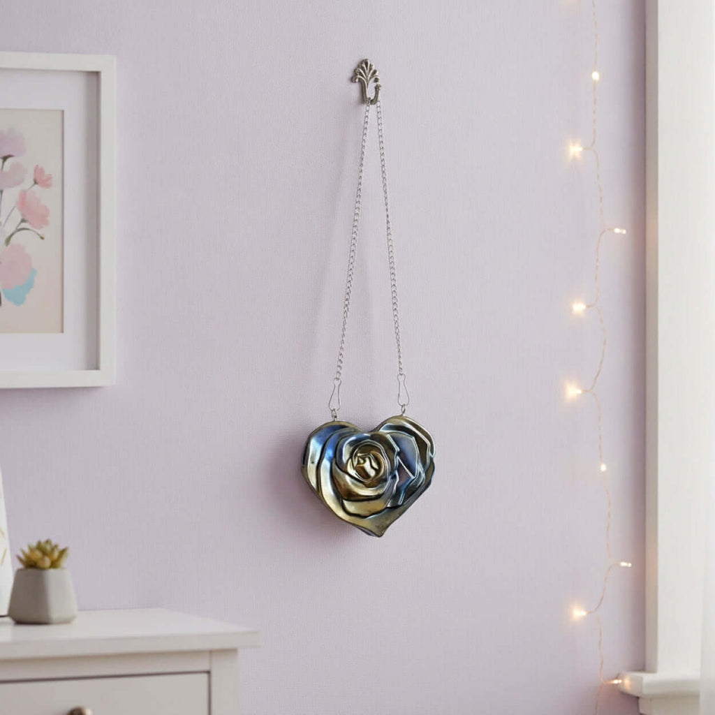 A metallic Heart Wall Decal hanging suspended from a hook on a light lavender wall, with a small white cabinet, framed art, and warm string lights visible in the room.