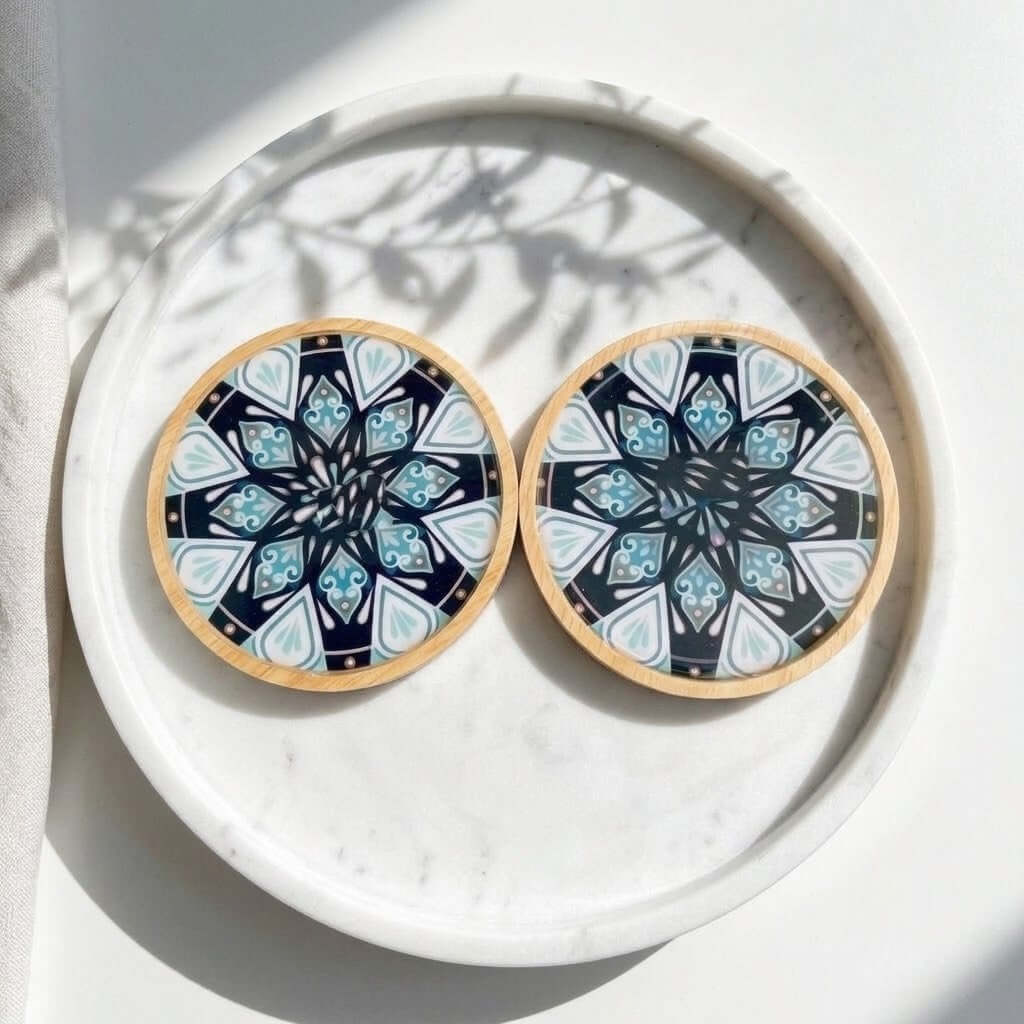 Mandala Bamboo Coasters - Two round coasters with a light wood rim and a black, white, and blue mandala pattern, displayed on a white marble tray with leaf shadows.