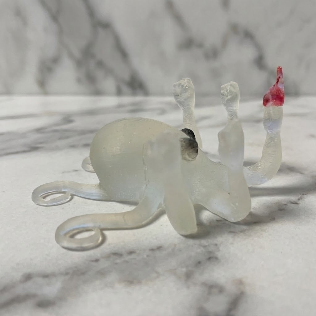 Novelty Octopus Figurine - A small translucent white octopus figurine with one raised arm ending in a red tipped finger, photographed on a light marble surface with a matching marble background.