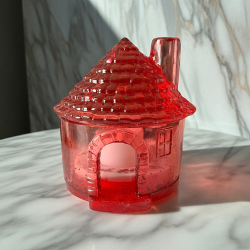 Candle Holder Houses - A translucent red house-shaped object with a textured conical roof and chimney, photographed on a grey marble surface with a matching marble background.