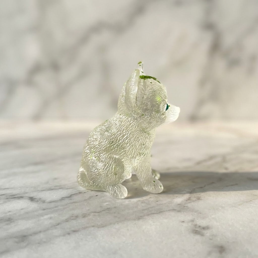 Chihuahua Dog Figurines - Side view of a small clear resin figurine shaped like a seated dog, with tiny green inclusions near the top, photographed on a white marble surface against a softly blurred marble background.