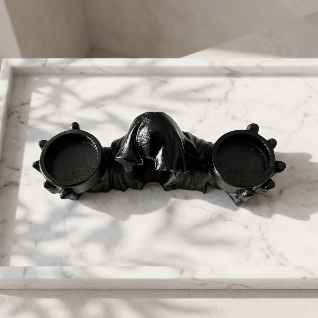Grim Reaper Candle Holder - Top view of a glossy black ceramic gothic-style candle holder with a central hooded figure flanked by two small candle cups, displayed on a white marble tray with dappled sunlight.