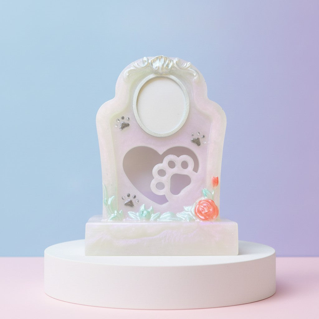 Pet Memorial A pastel memorial-style photo plaque or keepsake display on a round pedestal. The decorative white and pale pink plaque features an oval photo slot at the top, a heart-shaped cutout with a paw print silhouette in the center, small paw print and flower accents, and is set against a soft blue-to-lavender gradient background.