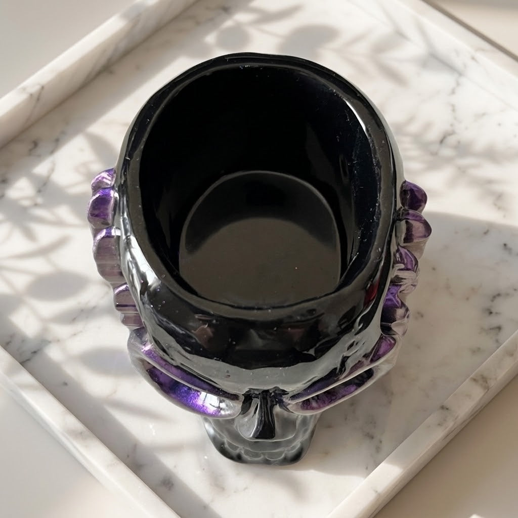 Resin Skull Pots - A glossy black skull-shaped cup or candle holder with purple crystal-like accents on the sides, photographed from above on a white marble tray.
