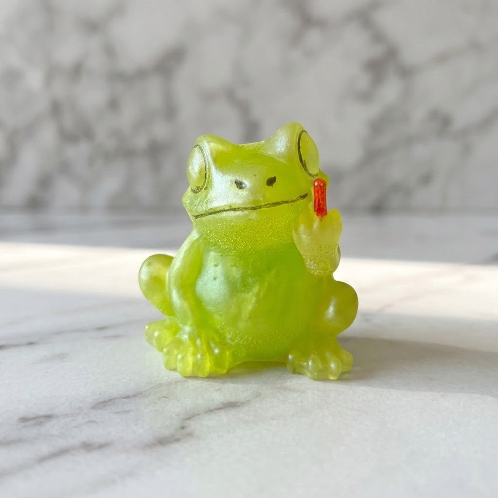 Novelty Frog Figurine - A small translucent lime-green frog figurine sitting on a white marble surface with a blurred grey marble background. The frog holds up a tiny red finger in one of its front paws.