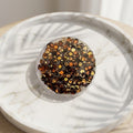 Golden Sequin Coasters - A round coaster filled with small metallic sequins in gold, bronze, and dark brown tones, placed on a white marble tray with leaf shadows cast across it.