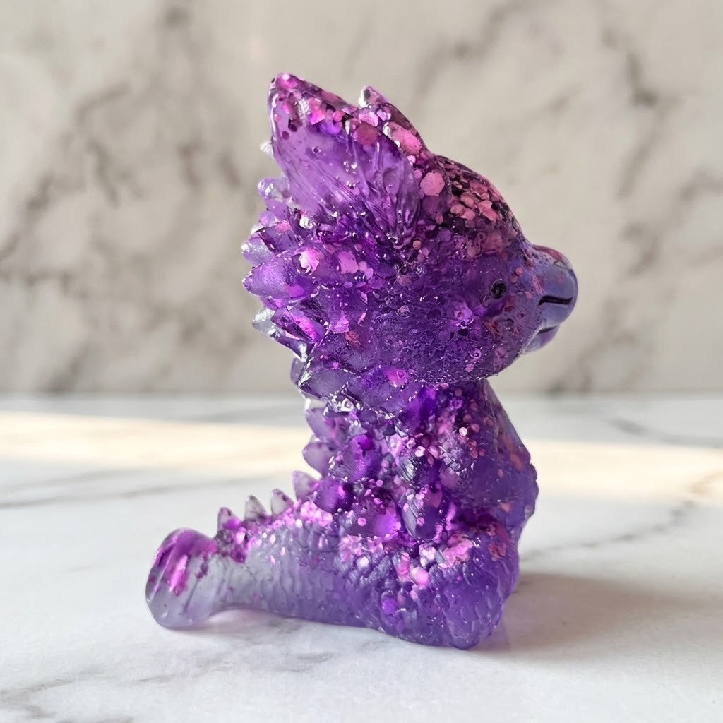 Baby Dragon Figurines - Side view of a small translucent purple dragon figurine with glittery pink and silver flecks, sitting on a white marble surface against a blurred grey marble background.