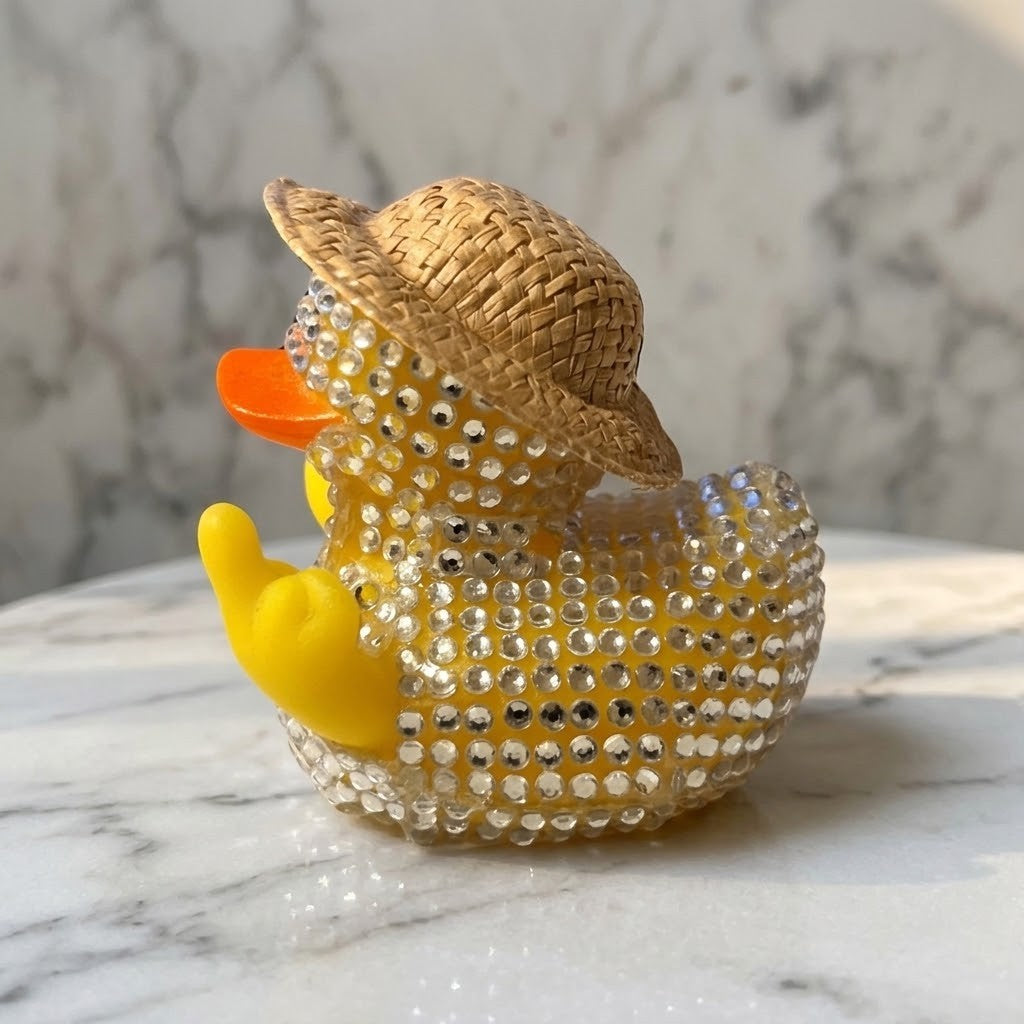 Diamante Duck Figurine - Side view of a yellow rubber duck styled with a woven straw hat and covered in clear rhinestones, sitting on a white marble surface with a marble-patterned background.