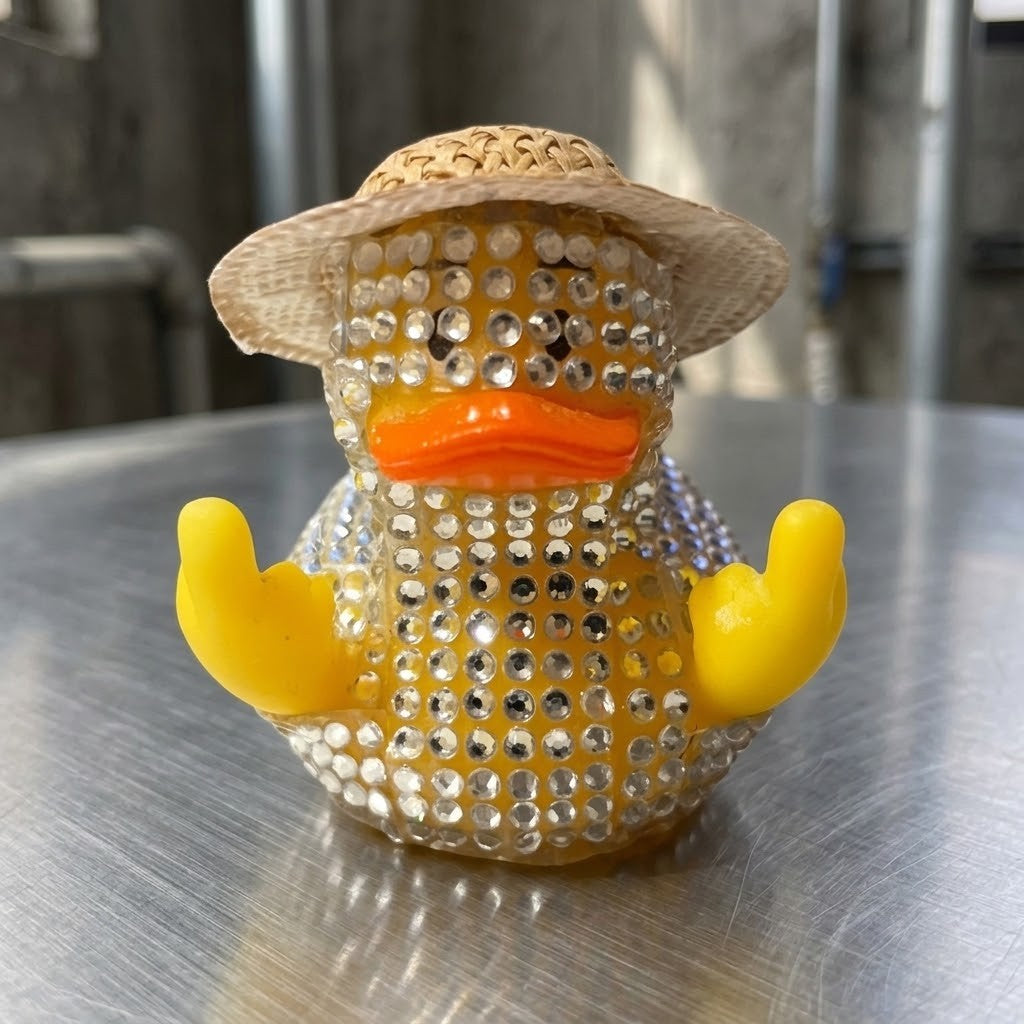Diamante Duck Figurine - A rhinestone-covered rubber duck toy wearing a straw hat, with a bright orange beak and yellow middle fingers pointing upwards, sitting on a brushed metal surface.