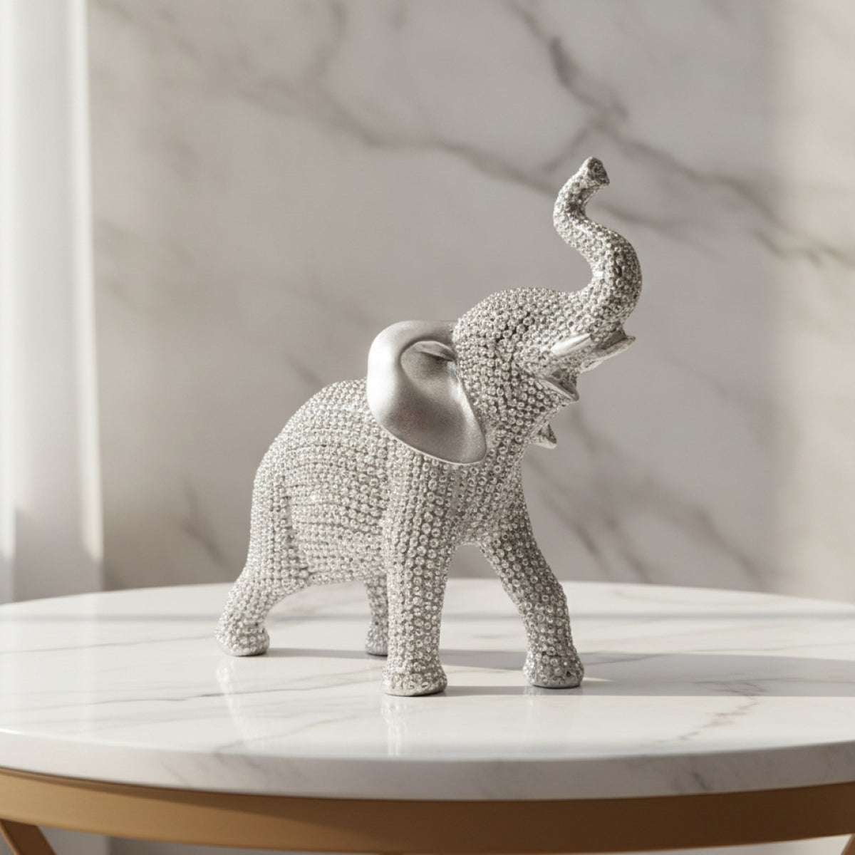 A rhinestone-embellished elephant figurine with a polished silver ear, standing on a white marble-topped table against a marble-patterned wall.