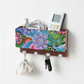 A wall-mounted Storage Rack Box wooden organizer with a colorful mandala-patterned front panel, silver hooks, and storage slots holding a charger, remote, and cord. A small fabric pouch and keys hang from the hooks against a plain white wall.