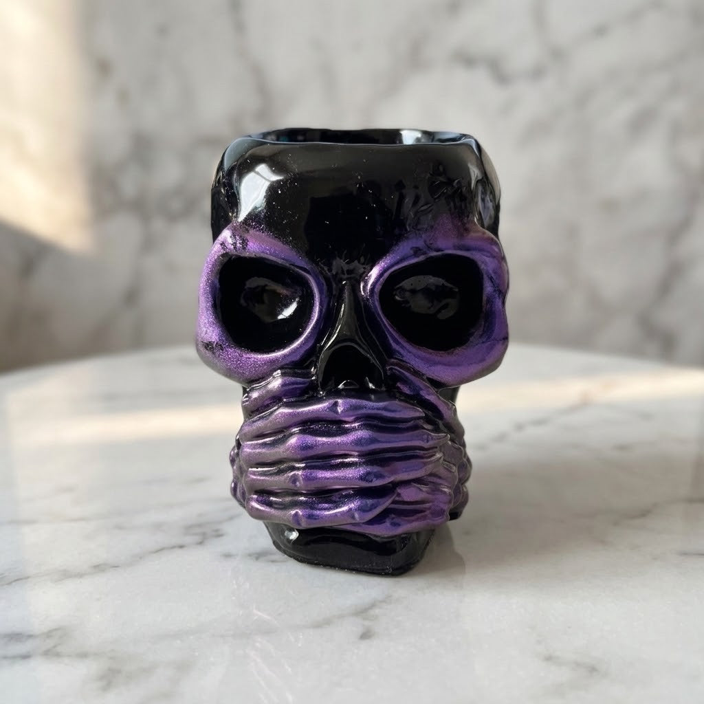 Resin Skull Pots - A glossy skull-shaped pot with a black and metallic purple glaze, set on a white marble surface against a matching marble background.