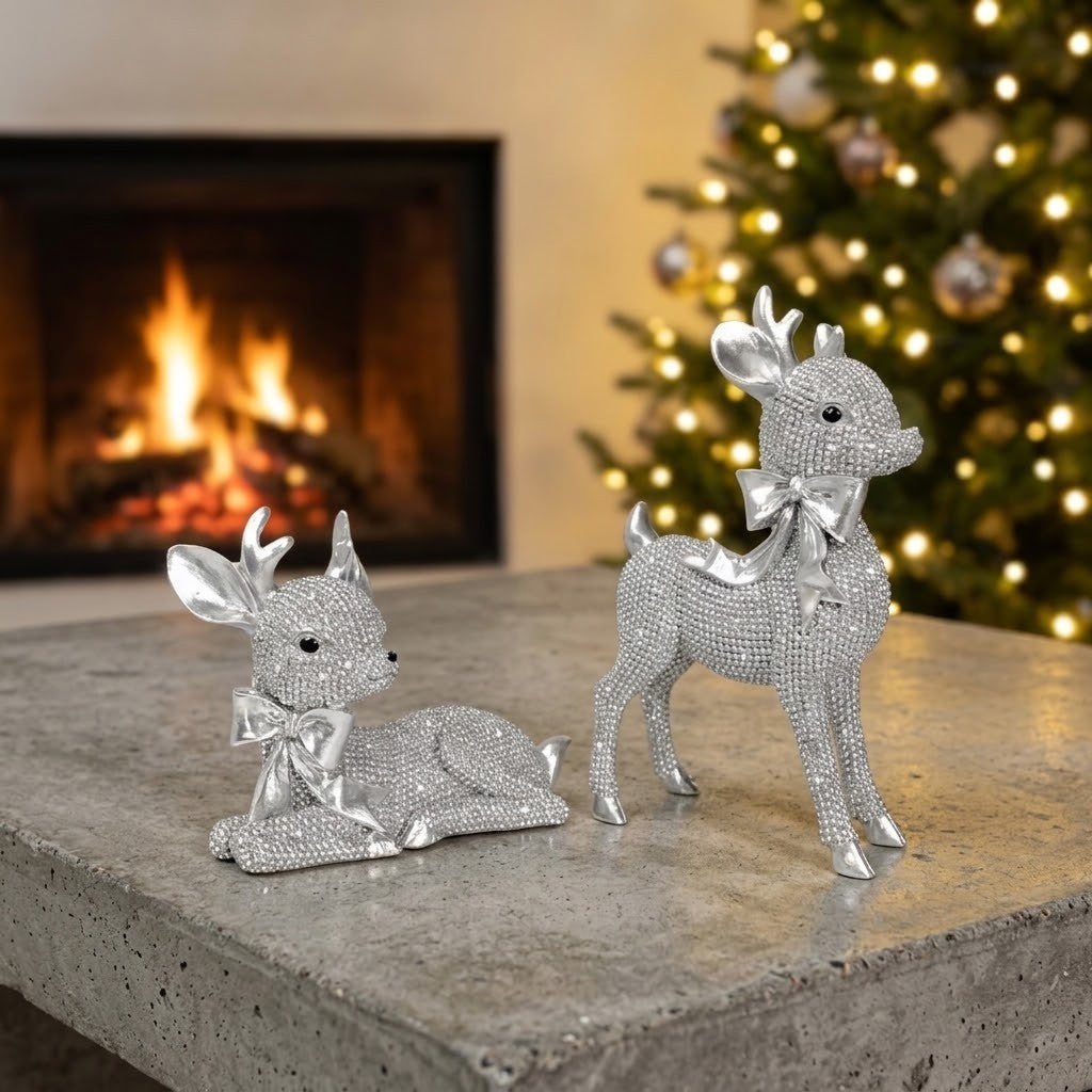 Reindeer Gift Set - Two silver rhinestone reindeer figurines, one lying down and one standing, displayed on a stone mantel with a warm fireplace and a lit Christmas tree in the background.