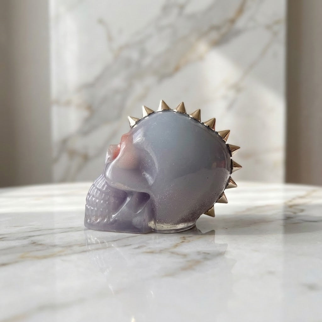 Studded Gothic Skull - Side view of a skull-shaped figurine with a glossy lavender-purple body and metallic silver spikes around the top, displayed on a white marble surface with a marble background. 