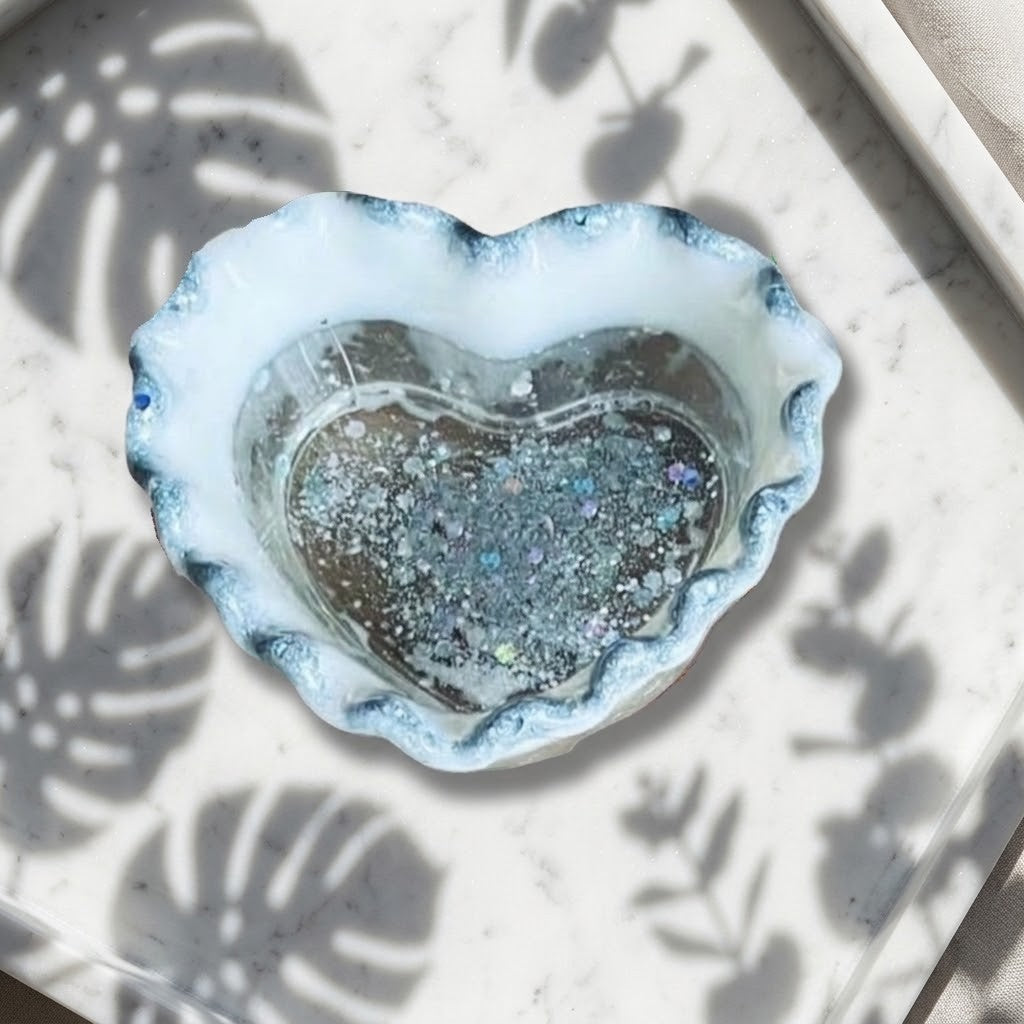 Heart Shaped Trinket Jar - A heart-shaped resin tray with a scalloped silver and white rim, containing glittery silver pieces, placed on a white marble surface with leaf shadows.