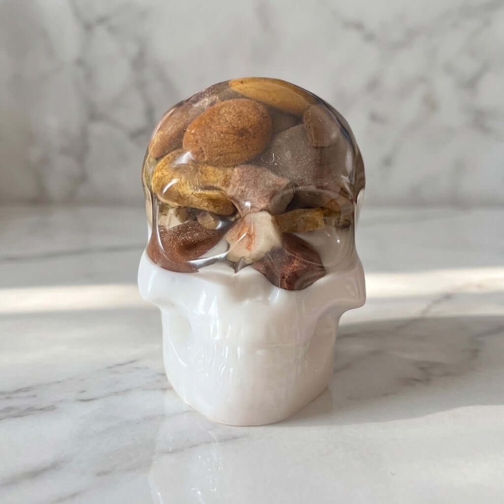 Gothic Resin Stone Skull - A decorative skull sculpture with a glossy transparent top section filled with polished stones and a solid white lower section, displayed on a white marble surface with a matching marble background.