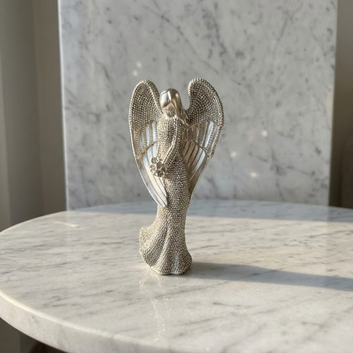 Christmas Angel Figurine - A silver-toned angel figurine with detailed textured wings and a flowing gown, displayed on a white marble table against a marble wall.