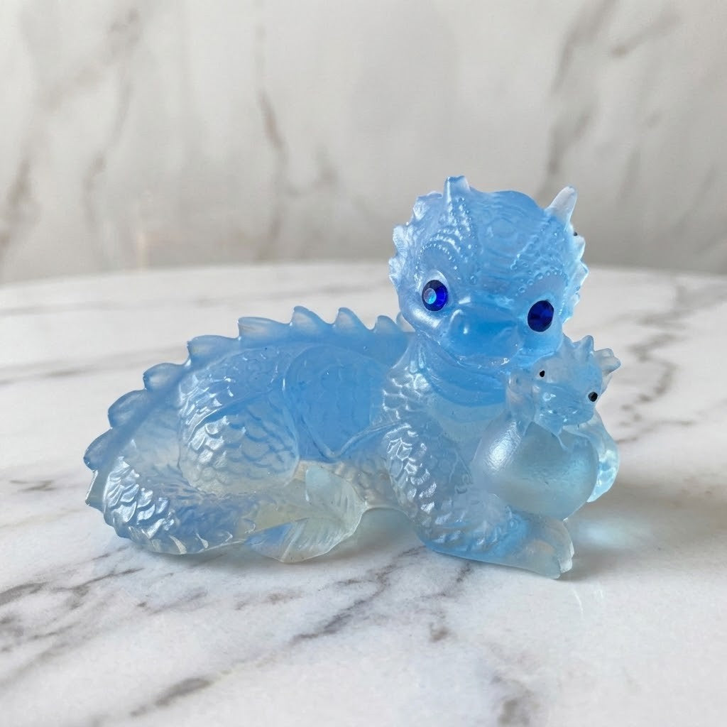 Dragon Figurines - A small transparent blue dragon figurine with dark blue eyes sits on a white marble surface, against a matching marble wall, holding a blue baby dragon and egg in its front paw