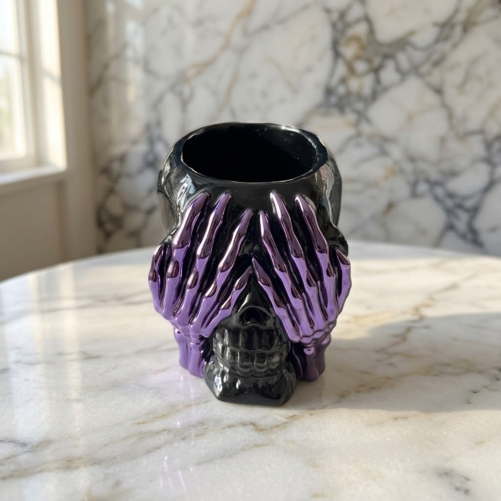 Resin Skull Pots - A skull-shaped pot with a glossy black finish, featuring skeletal hands covering the eyes in a metallic purple colour, sitting on a white marble table with a marble wall background.