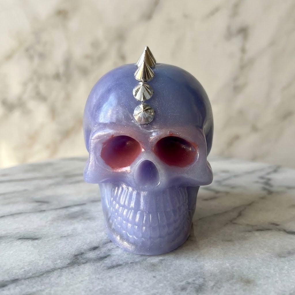 Studded Gothic Skull - A small skull-shaped decorative object in a lavender-purple glossy finish and red eyes, with metallic silver spikes on top, photographed on a marble surface against a softly blurred marble background.