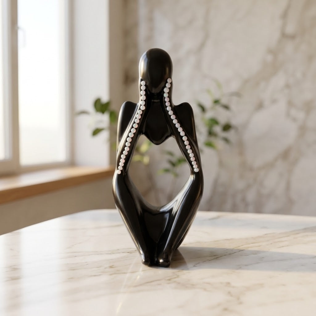 Nordic Resin Thinker - Front view of a glossy black abstract figurative sculpture of a seated female form with white pearl-like accents along the arms, displayed on a light marble surface in a bright interior setting.