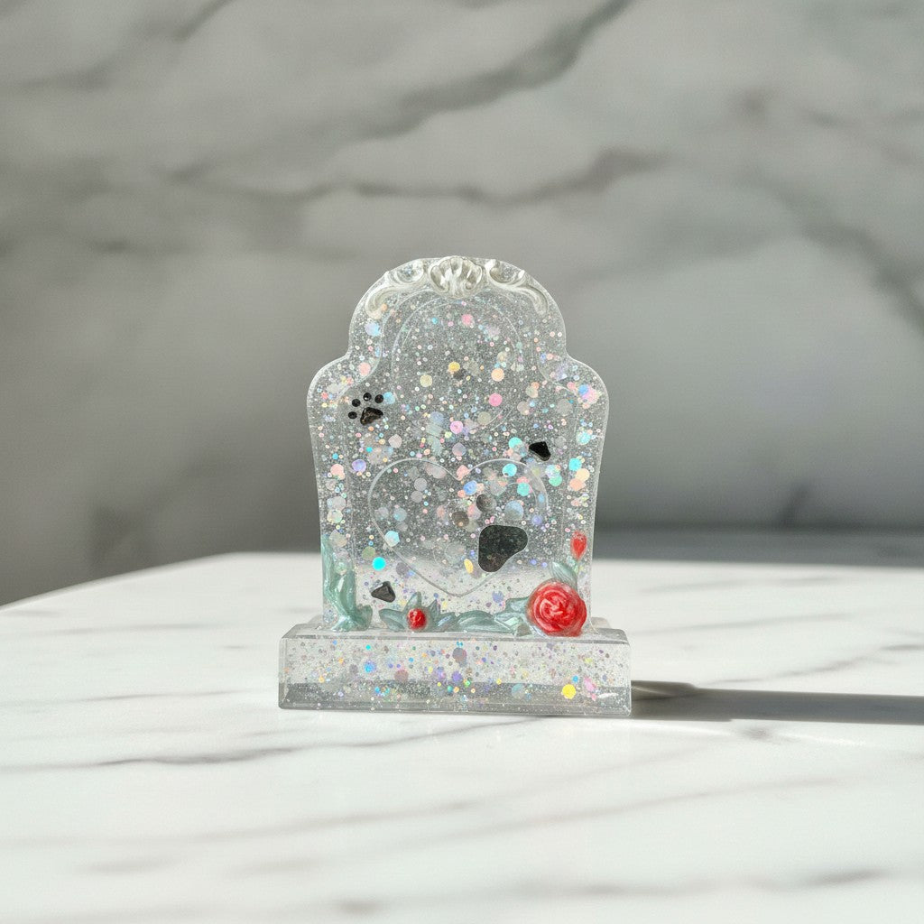 Pet Memorial - A small clear resin tombstone-shaped decorative object filled with iridescent glitter, small paw prints, and a red rose, displayed on a white marble surface against a dark marble wall