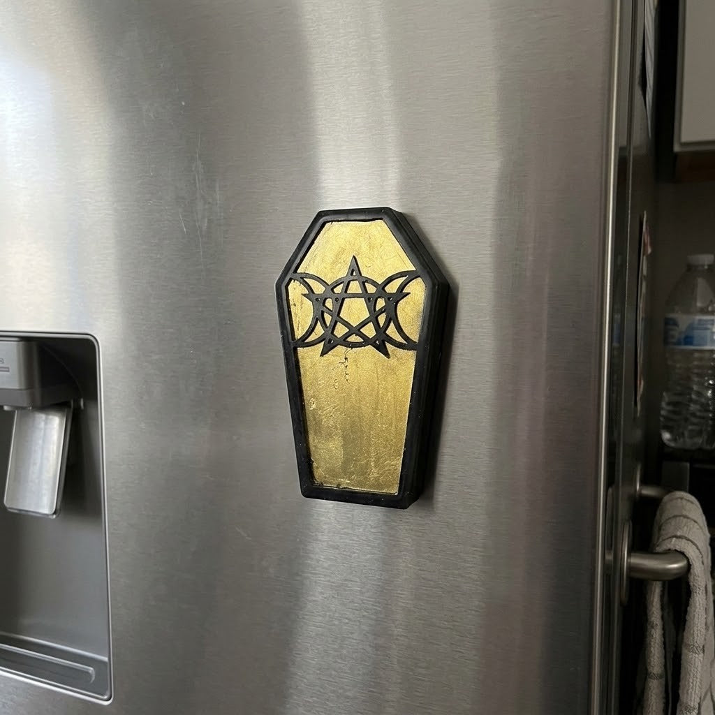 Coffin Shaped Fridge Magnets - A coffin-shaped refrigerator magnet with a black pentagram and triple moon symbol over a gold background, shown attached to a stainless steel refrigerator.
