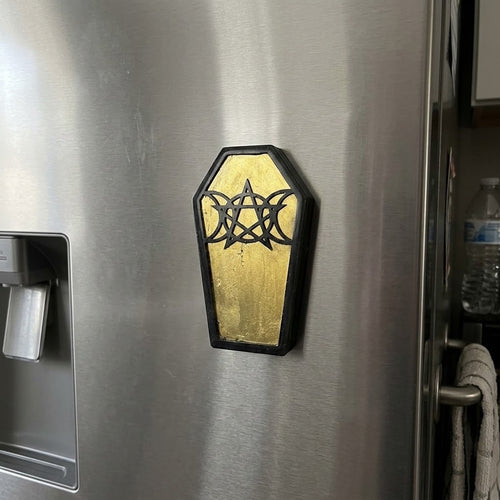 Coffin Shaped Fridge Magnets - A coffin-shaped refrigerator magnet with a black pentagram and triple moon symbol over a gold background, shown attached to a stainless steel refrigerator.