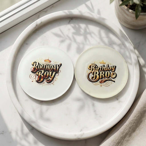 Birthday Boy Coasters - Two round white coasters with decorative 'Birthday Boy' and 'Birthday Bros' text designs, displayed on a white marble tray on a sunlit tabletop.