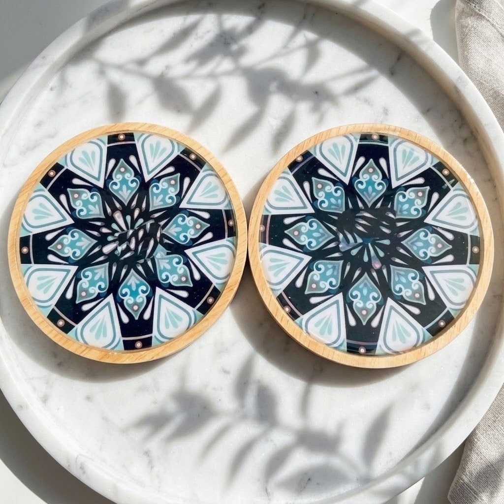 Mandala Bamboo Coasters - Two round coasters with a light wood rim and a black, white, and blue mandala pattern, displayed on a white marble tray with leaf shadows.